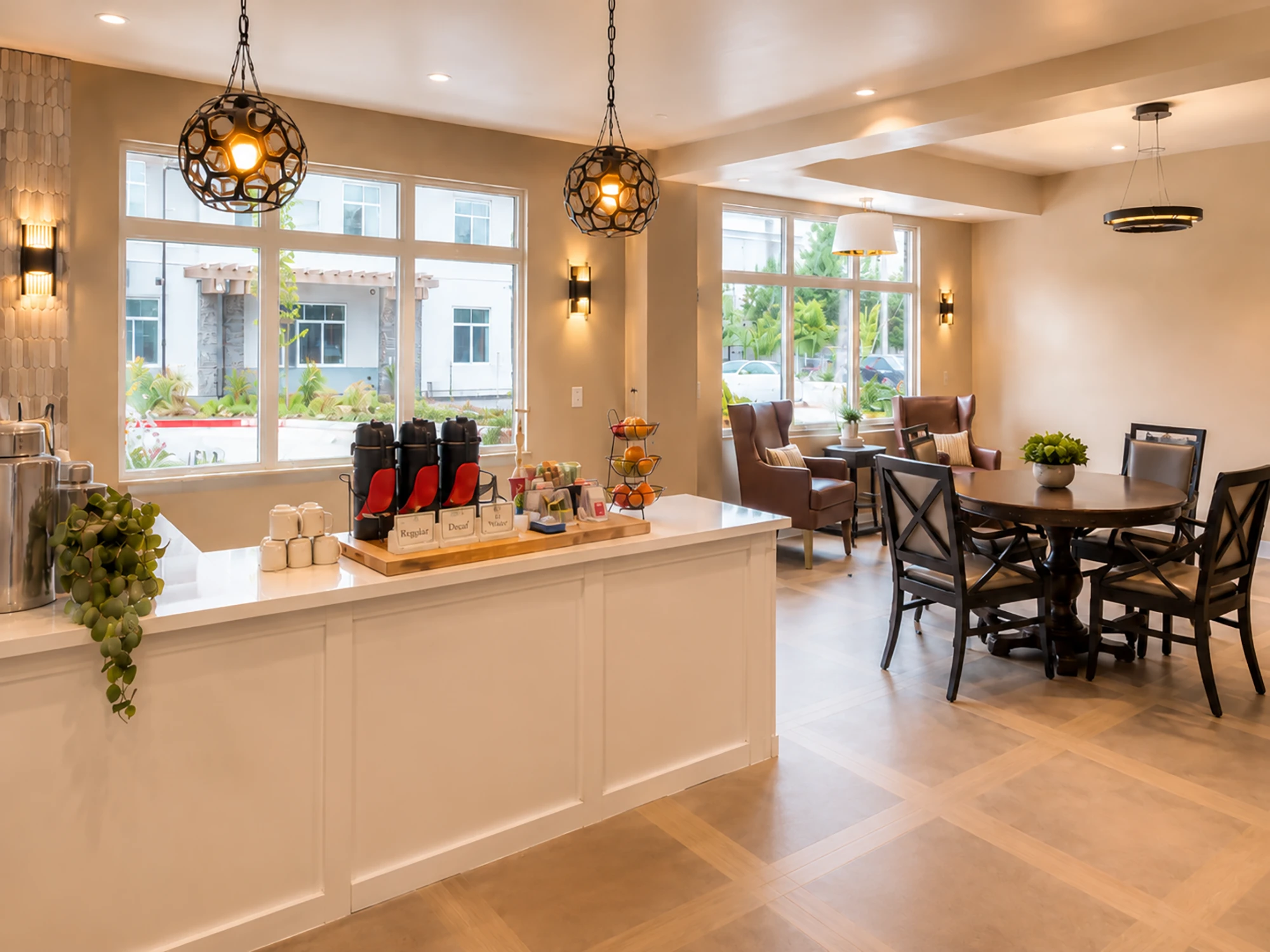 Cozy lounge area with armchairs, dining table, coffee dispensers, fruit stand, pendant lights, and large windows allowing natural light.